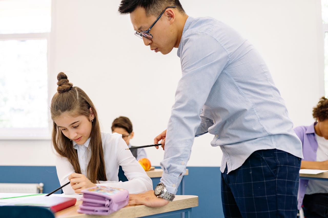 team-02 Asian male teacher assisting a young caucasian girl with her studies in a classroom setting.